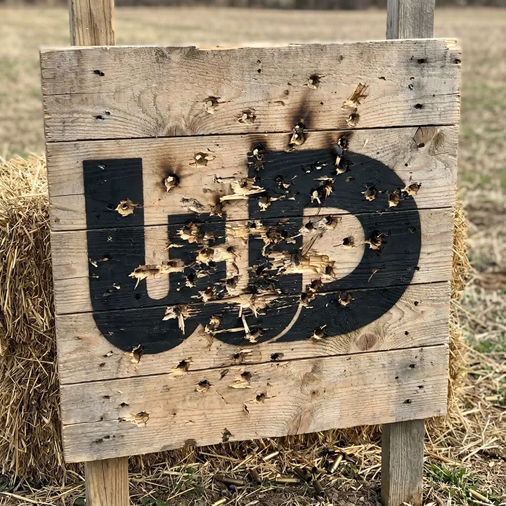 Wooden target board with a painted black Womp Designs logo heavily riddled with bullet holes.