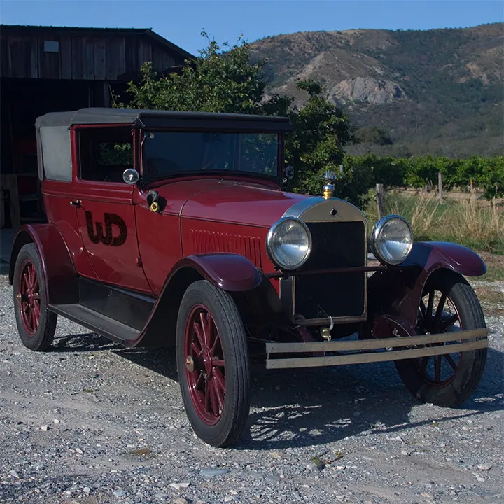 Vintage maroon antique car with a Womp Designs logo parked on gravel near a barn.