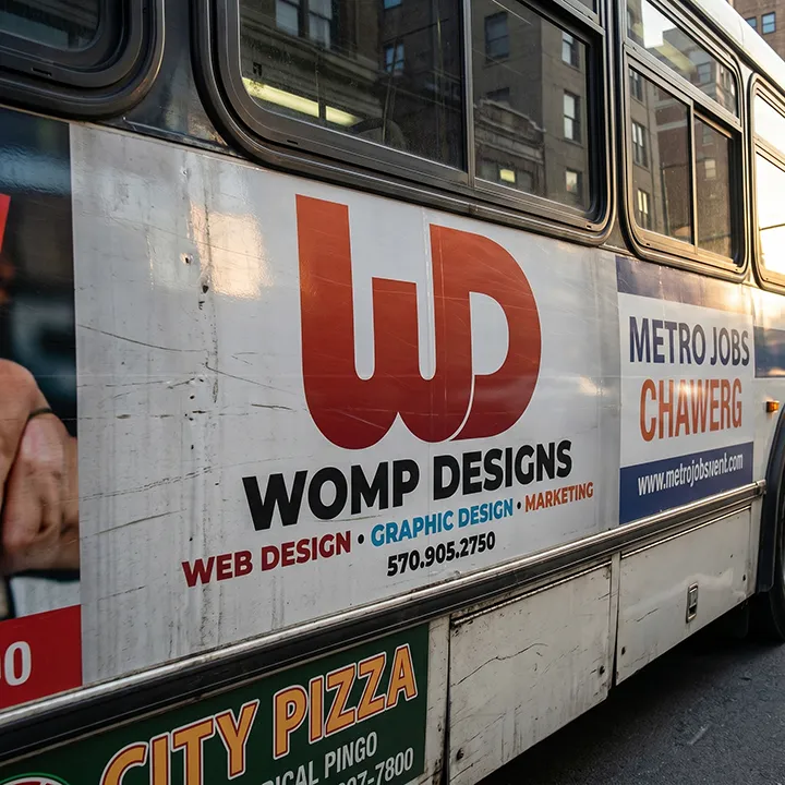 Side of a city bus showing a Womp Designs advertisement banner.