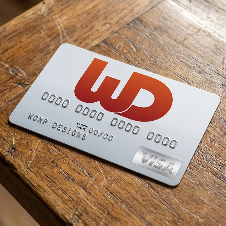 A silver Visa credit card with a large red-and-orange gradient WD logo, resting on a textured wooden surface. The name WOMP DESIGNS is embossed on the bottom left.